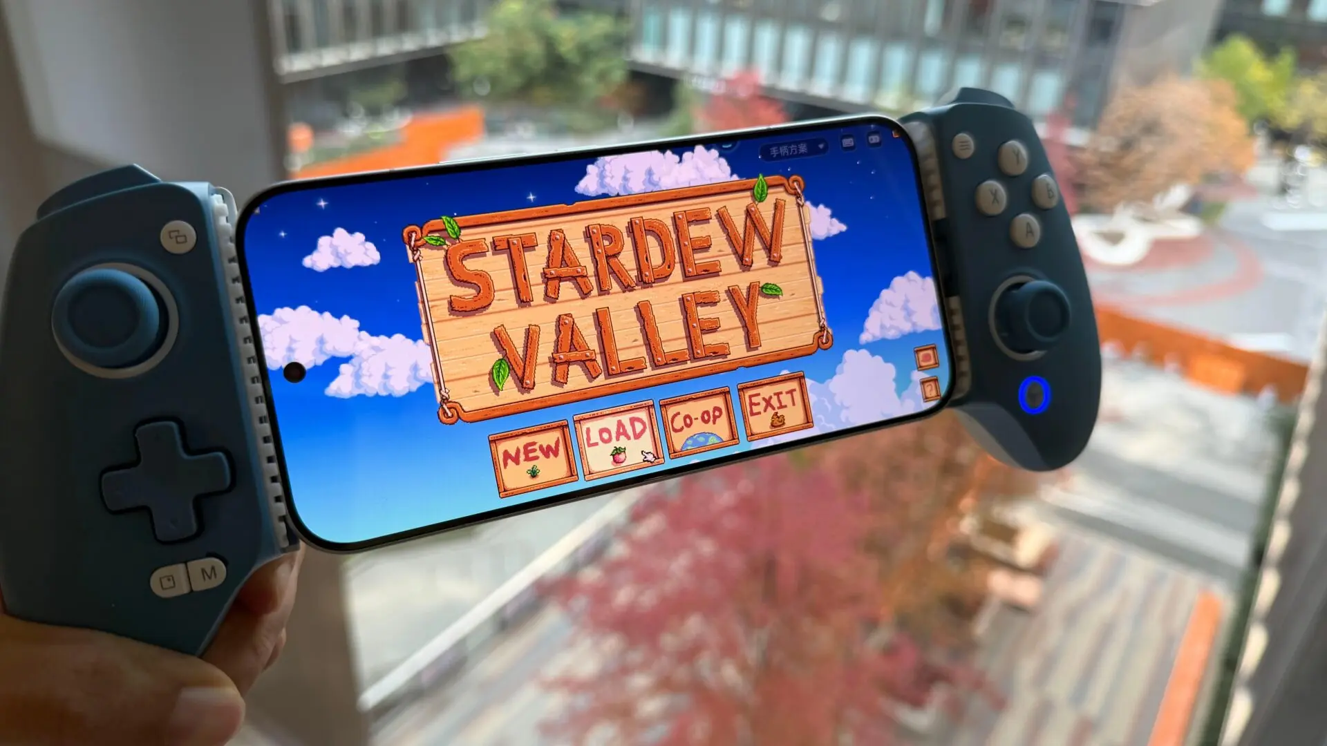 Смартфон с подключённым геймпадом, на экране запущена игра Stardew Valley, на фоне окно с видом на город